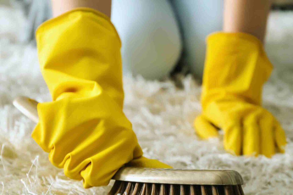 A person wearing yellow gloves scrubs a rug with a brush, focusing on cleaning and maintaining its appearance.