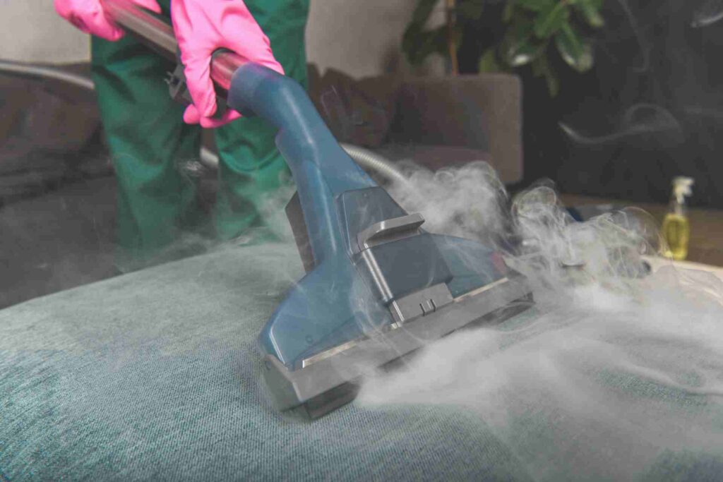 A person wearing gloves uses a steam cleaner to sanitize a couch.